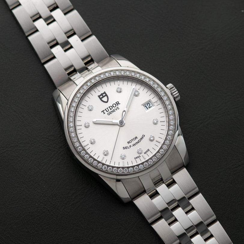 TUDOR Glamour Date silver wristwatch with white dial, date display, and diamond hour markers on metal bracelet, shown against a black background.