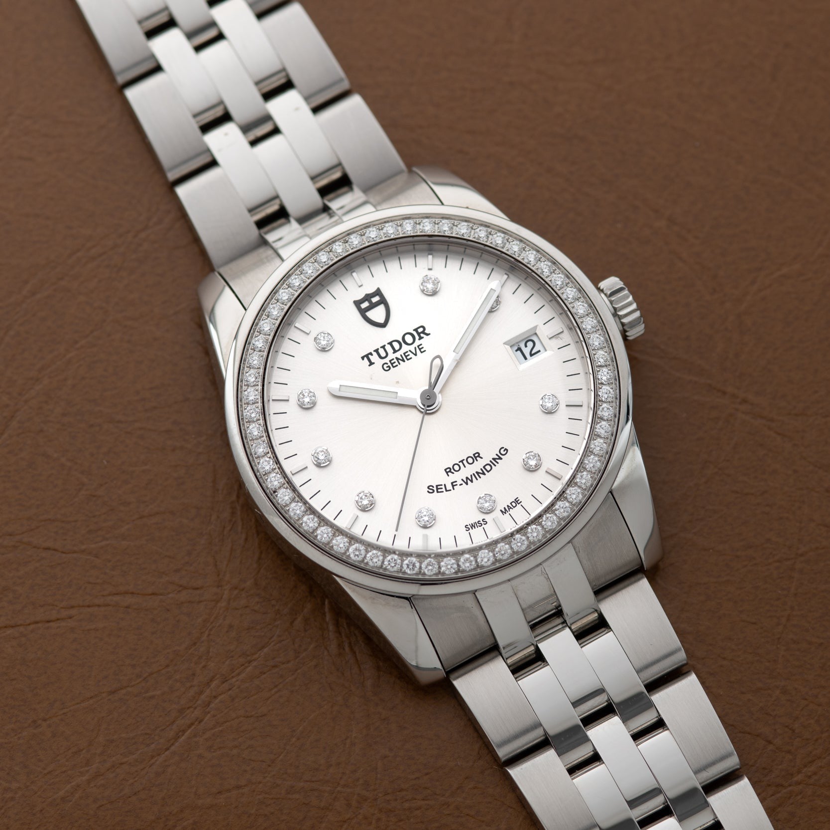 A TUDOR Glamour Date wristwatch with a diamond bezel rests on a brown leather surface.