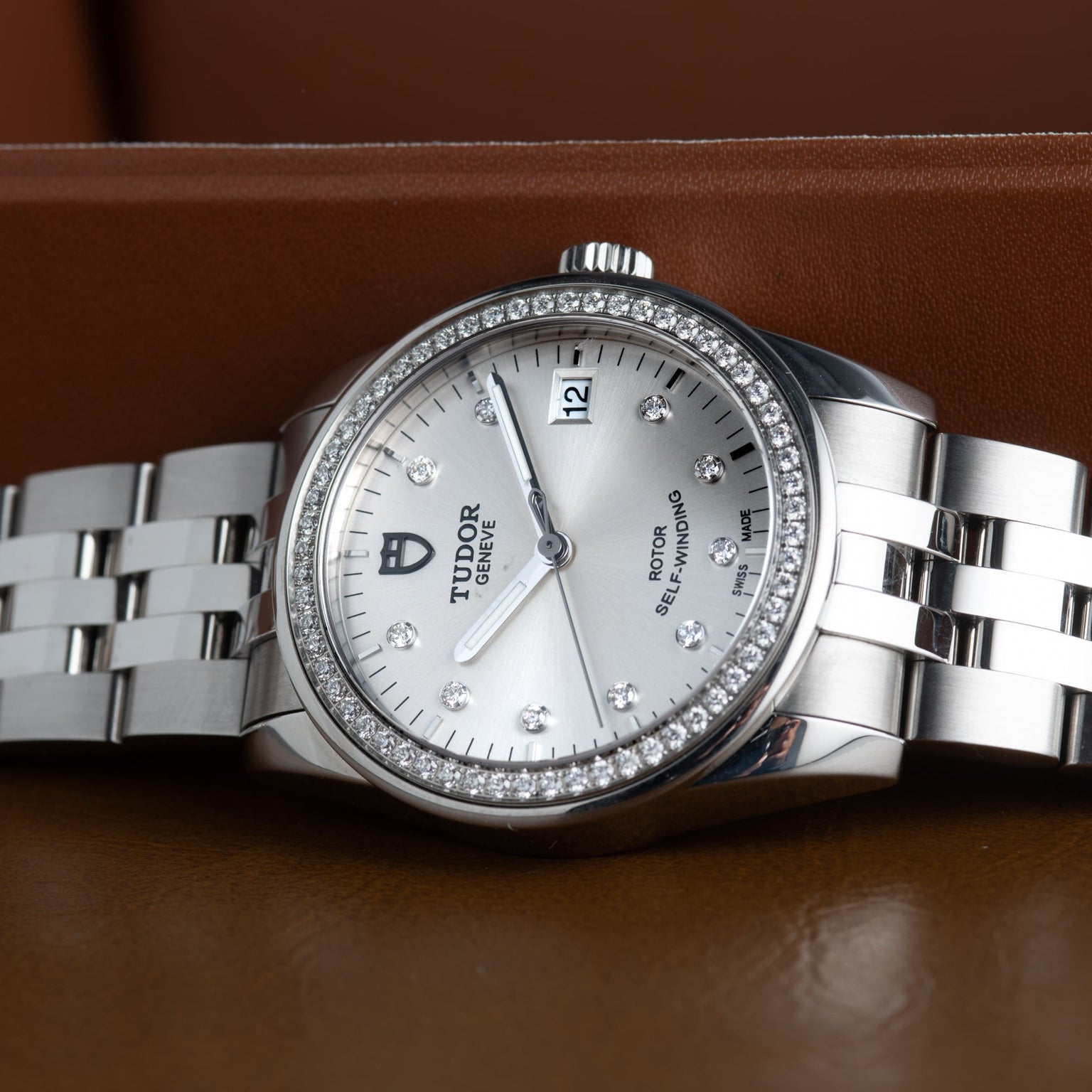 A TUDOR Glamour Date silver wristwatch with diamond bezel and markers, showcased on a brown leather surface.