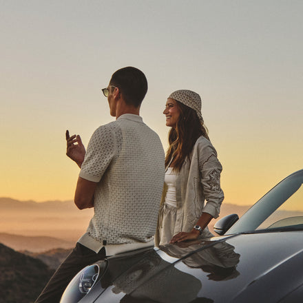 Two people lean on a car at sunset, enjoying the view and each others company in casual, stylish outfits.