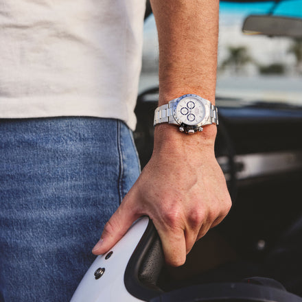 A person wearing a silver wristwatch and holding a white helmet, standing next to a car.