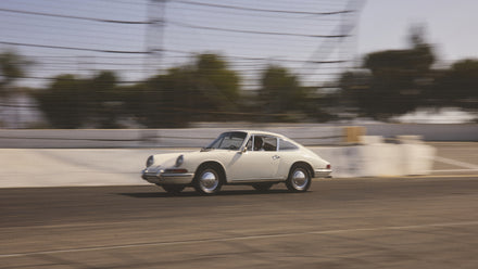 A vintage white sports car driving quickly on a racetrack with blurred trees in the background.