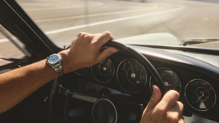 Close-up of hands on a vintage car steering wheel, wearing a silver watch, driving on a sunny day.