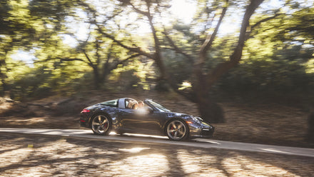 A black convertible sports car driving on a sunlit forest road with trees surrounding it.