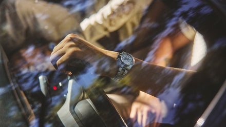 Close-up of a hand wearing a silver watch, gripping a steering wheel, seen through a car window with reflections.