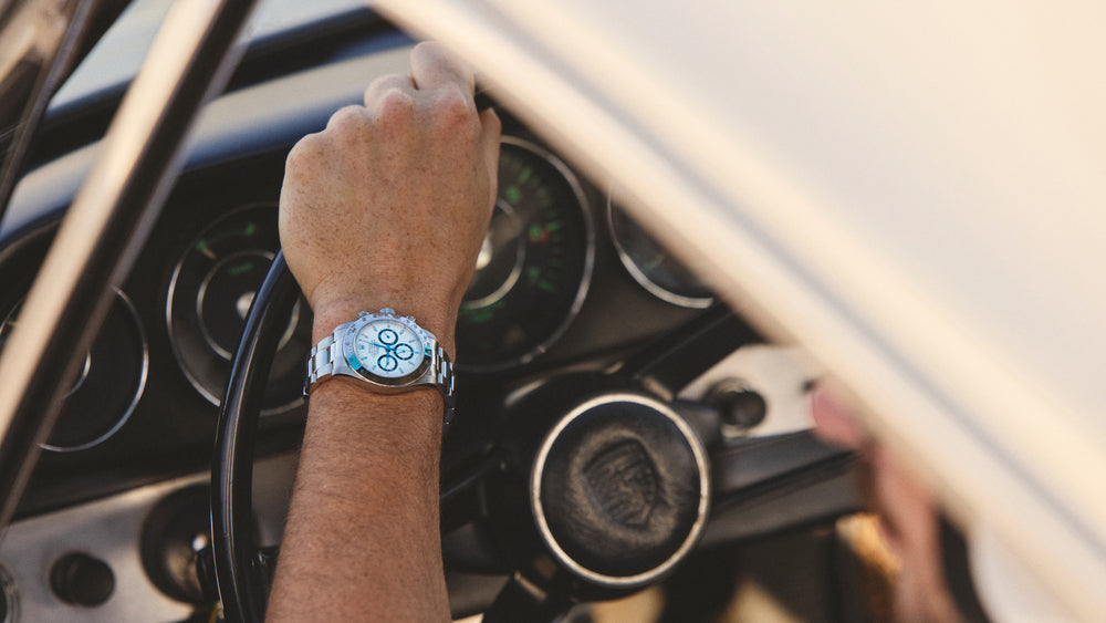 A hand grasping a steering wheel, the wrist wears a Rolex Daytona watch