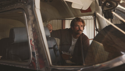 A man with a mustache sits inside a small aircraft, looking out the window while holding a map.
