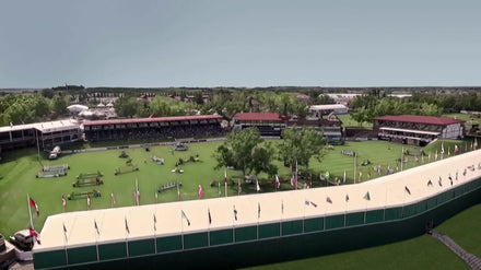 A large outdoor equestrian arena with jumps, surrounded by stands and flags on a sunny day.