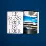 An open HODINKEE Magazine, Volume 12 by Hodinkee shows Le Mans: Hour by Hour on the left page and a blurred racetrack photo with classic cars under clouds on the right, reflecting vintage racing and watch culture.