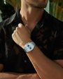 A man outdoors amid greenery wears a black sheer shirt and a Glashütte Original Seventies Chronograph Panorama Date wristwatch.