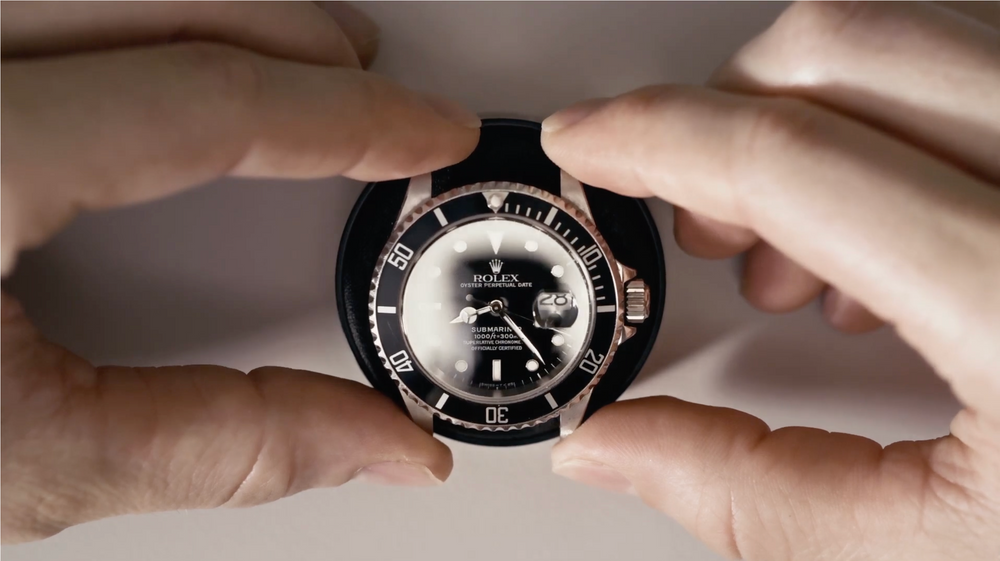 Two hands holding a black Rolex watch face with a silver bezel against a light background.