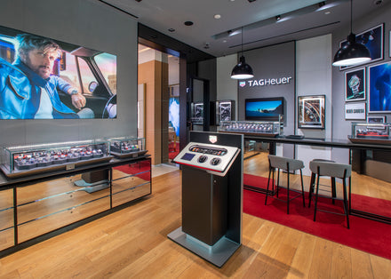 Modern TAG Heuer watch store interior with display cases, red carpet, and promotional posters on the walls.
