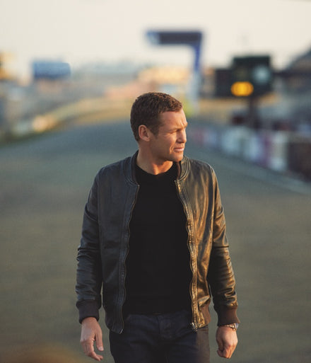 A man in a leather jacket stands outdoors on a road, looking to his left in soft, warm light.