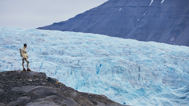 A man stands on a grey rock peak with a glacier in the background, behind the glacier is a mountain with streaks of snow