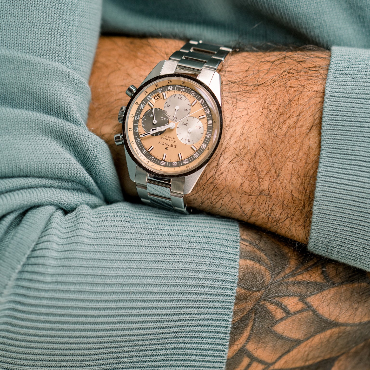 A person with a forearm tattoo wears the Zenith Chronomaster Original Limited Edition For Hodinkee, featuring a beige and silver dial, paired with a light blue long-sleeve top, highlighting its classic Zenith design.