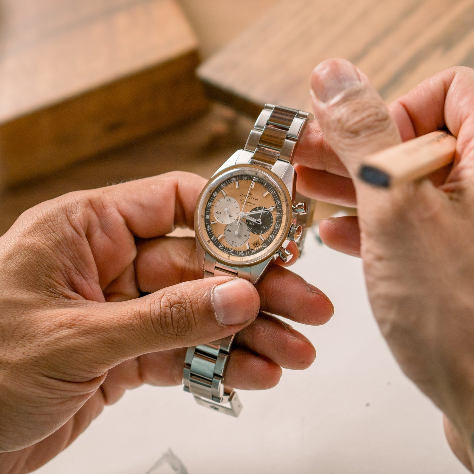 A person holds the Zenith Chronomaster Original Limited Edition For Hodinkee, a silver wristwatch with a brown and silver face, touching it with a wooden tool.