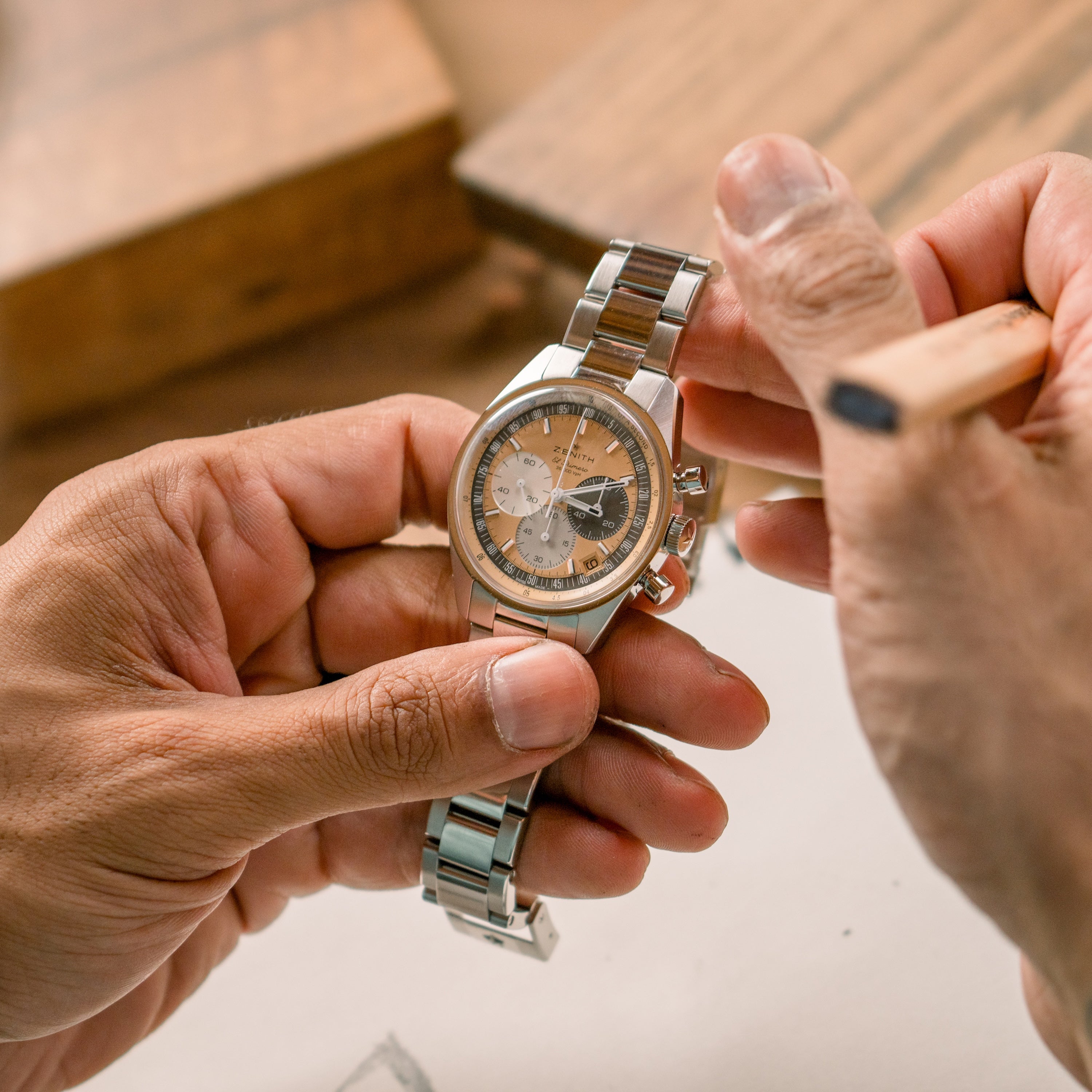 A person holds the Zenith Chronomaster Original Limited Edition For Hodinkee, a silver wristwatch with a brown and silver face, touching it with a wooden tool.