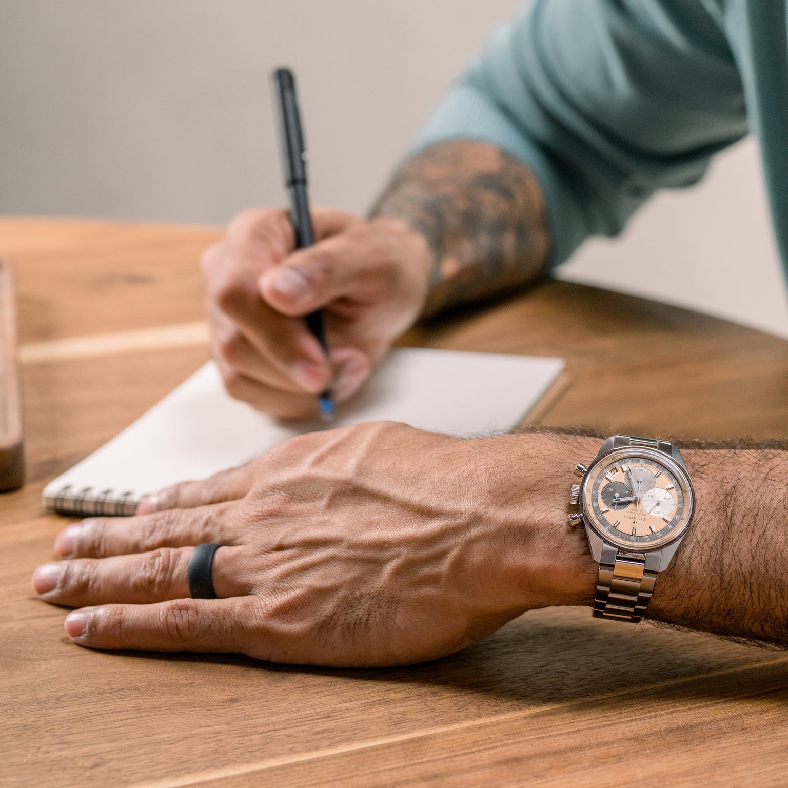A person wearing a Zenith Chronomaster Original Limited Edition For Hodinkee watch and a black ring writes in a notebook on a wooden table.
