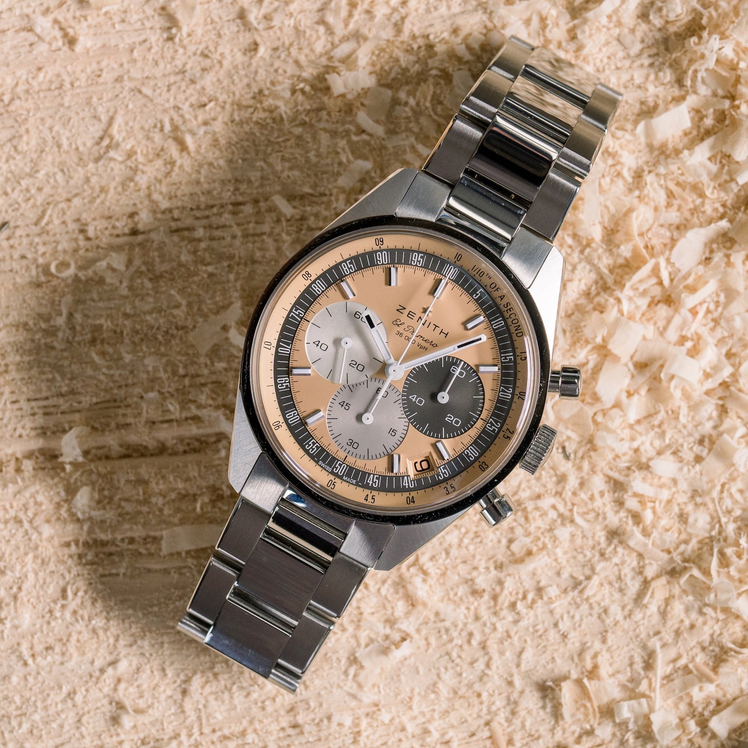 The Zenith Chronomaster Original Limited Edition For Hodinkee, with a stainless steel case and a multi-toned dial featuring three subdials, is displayed on a textured wooden surface scattered with wood shavings.