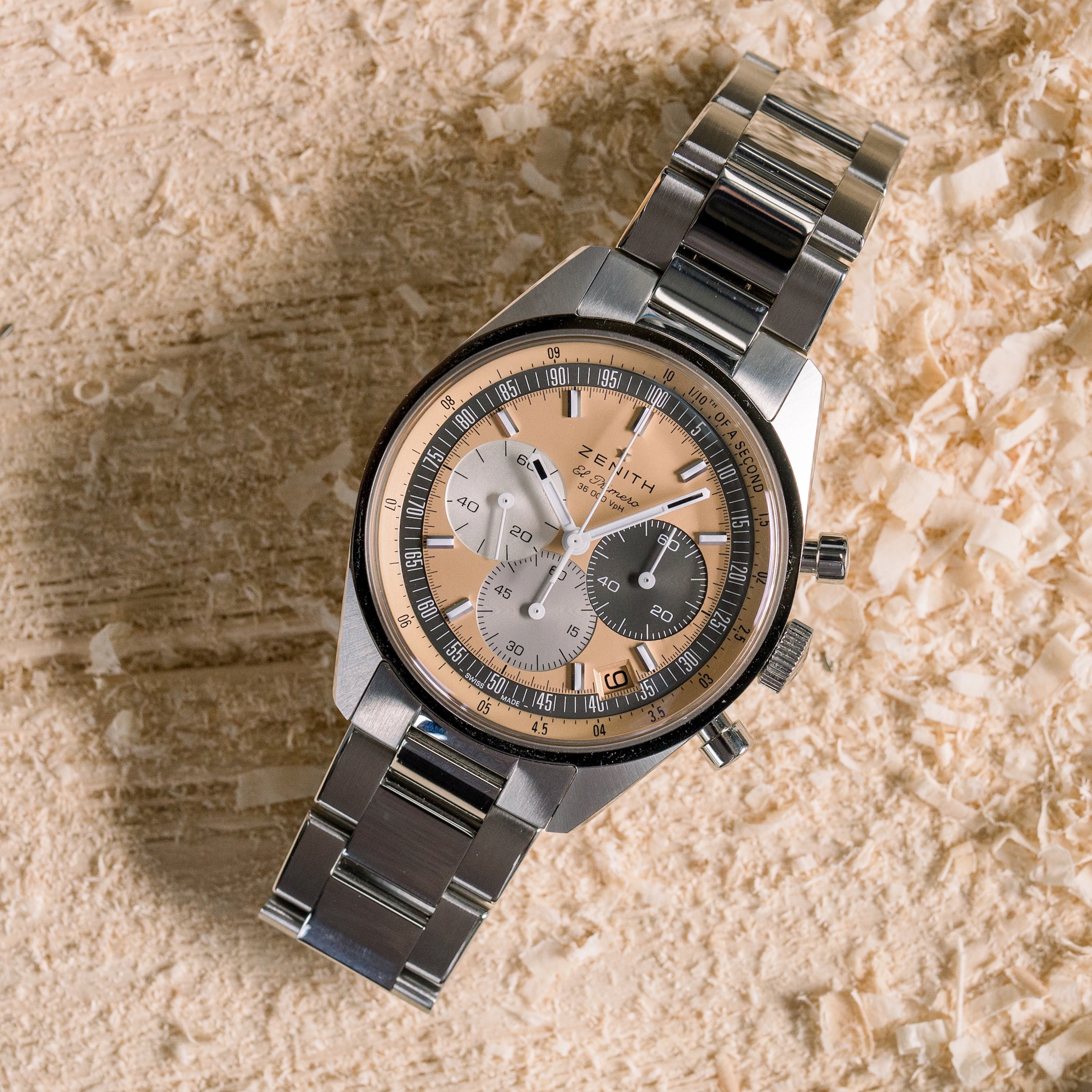 The Zenith Chronomaster Original Limited Edition For Hodinkee, with a stainless steel case and a multi-toned dial featuring three subdials, is displayed on a textured wooden surface scattered with wood shavings.