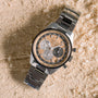 The Zenith Chronomaster Original Limited Edition For Hodinkee, with a stainless steel case and a multi-toned dial featuring three subdials, is displayed on a textured wooden surface scattered with wood shavings.