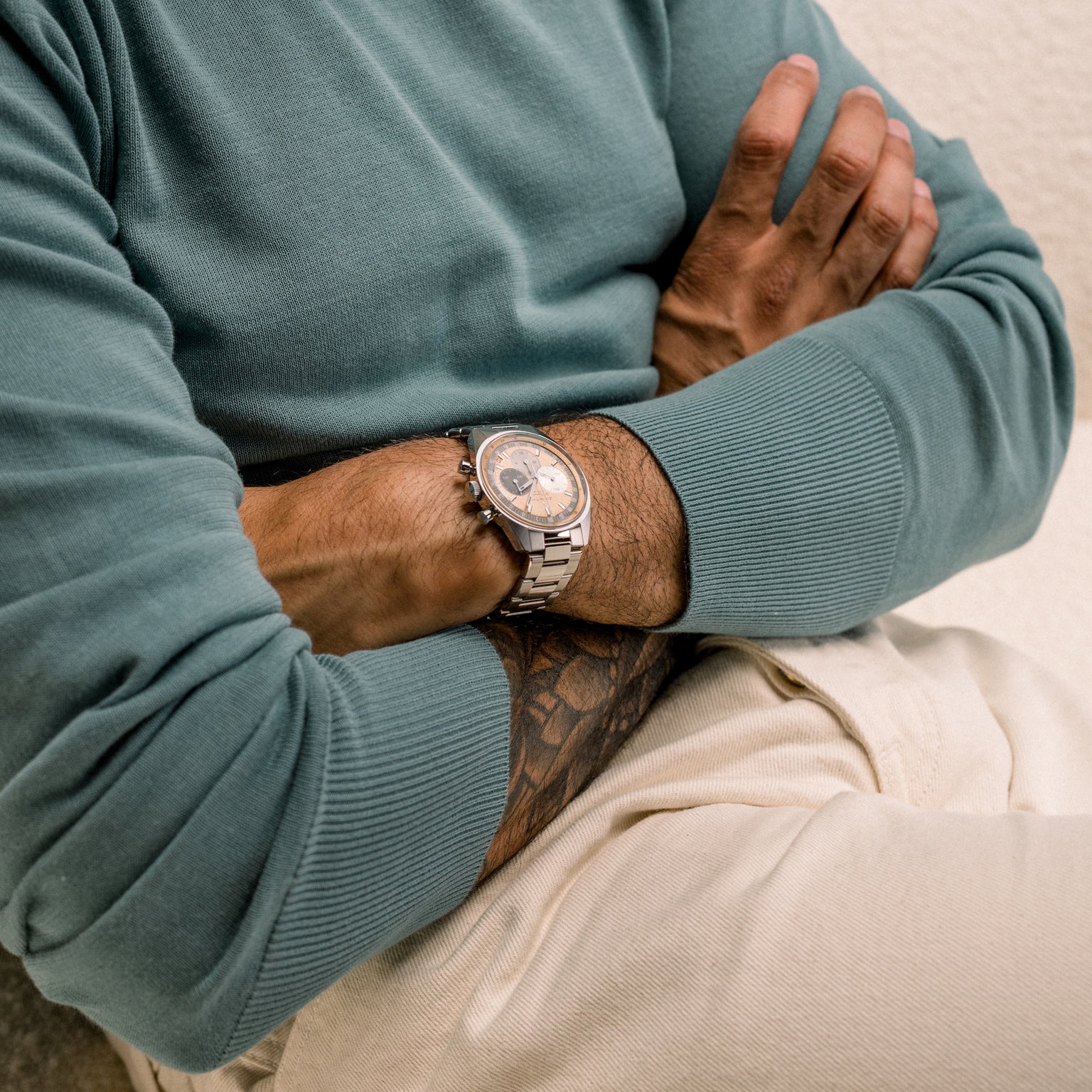 A person in a green sweater with crossed arms, wearing beige pants and a Zenith Chronomaster Original Limited Edition For Hodinkee watch by Zenith.