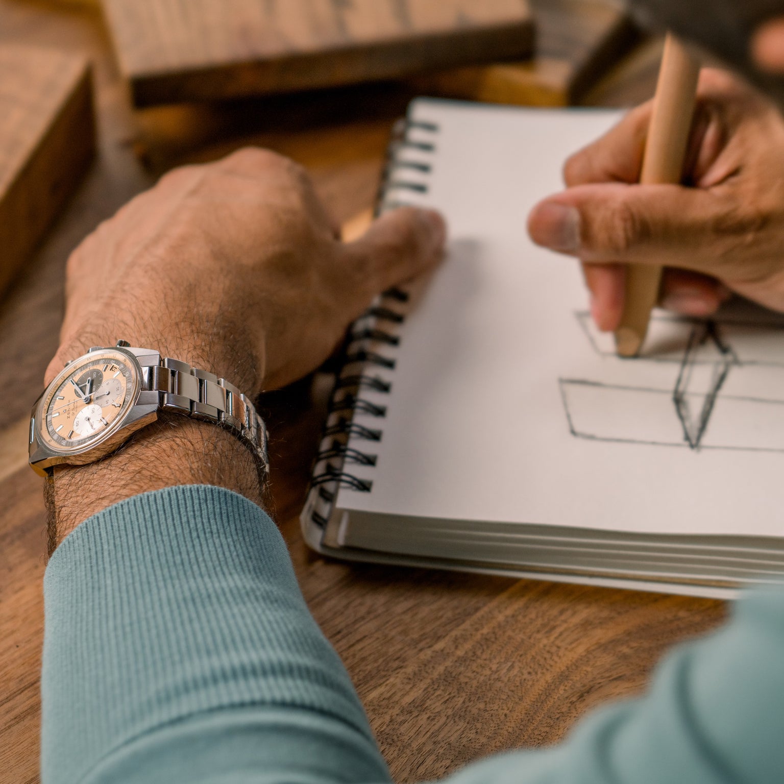 Wearing a Zenith Chronomaster Original Limited Edition For Hodinkee, a person sketches a 3D geometric shape in a spiral notebook with pencil on a wooden table, reflecting creativity and Zenith’s timeless design.
