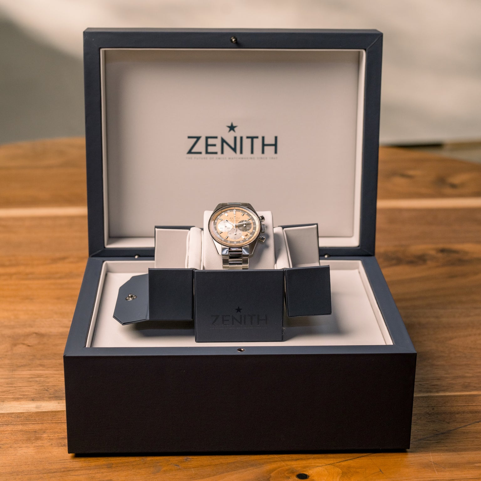 A Zenith Chronomaster Original Limited Edition For Hodinkee wristwatch with a classic design and silver band is displayed in an open black and white Zenith-branded box on a wooden surface.
