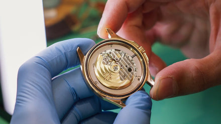 Person wearing blue gloves holding a gold watch mechanism for inspection.