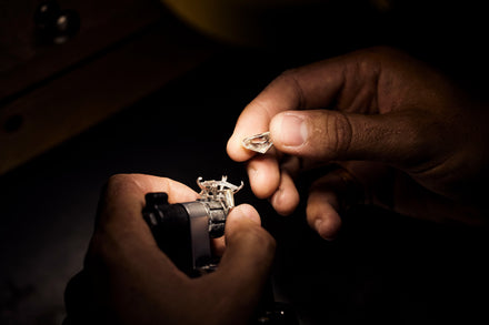 A person's hand holds a cut gem stone to place it into the prongs of a ring held in a jeweler's vice