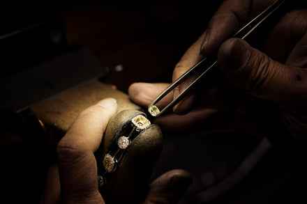 A jewelry clamp holds a jeweled setting as a person's hand with tweezers sets a yellow stone into place in the setting