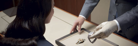 A person presents luxury watches on a tray to a seated woman in a store, wearing gloves.