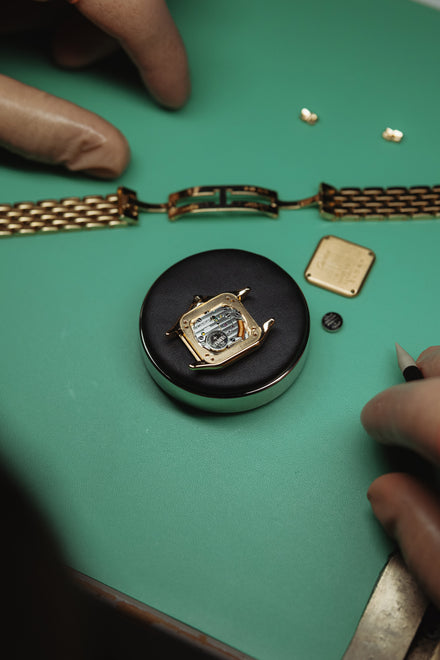 Hands assembling a gold wristwatch with tools and parts on a green work surface.