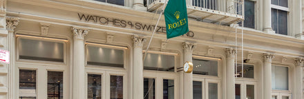 Storefront of Watches of Switzerland with a green Rolex flag and gold Rolex clock outside.