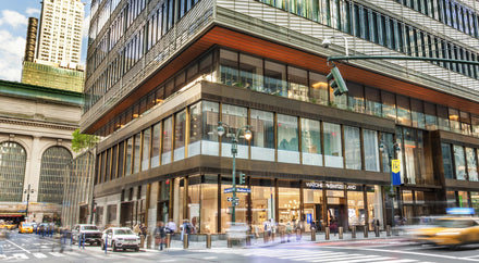 A busy city street corner with modern glass buildings and pedestrians crossing.
