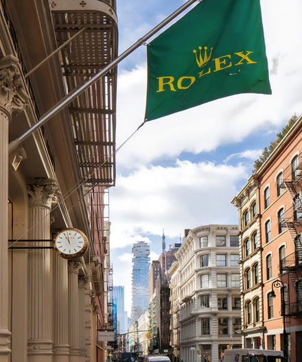 A green Rolex flag and a clock hang outside a building on a city street with historic architecture.