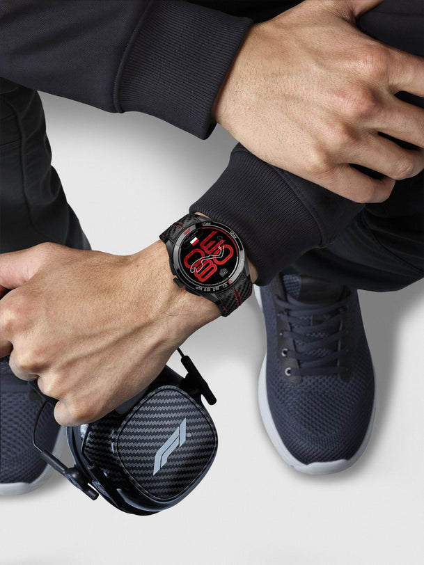 Person wearing a smartwatch with a black case and red display, sitting on a white surface.