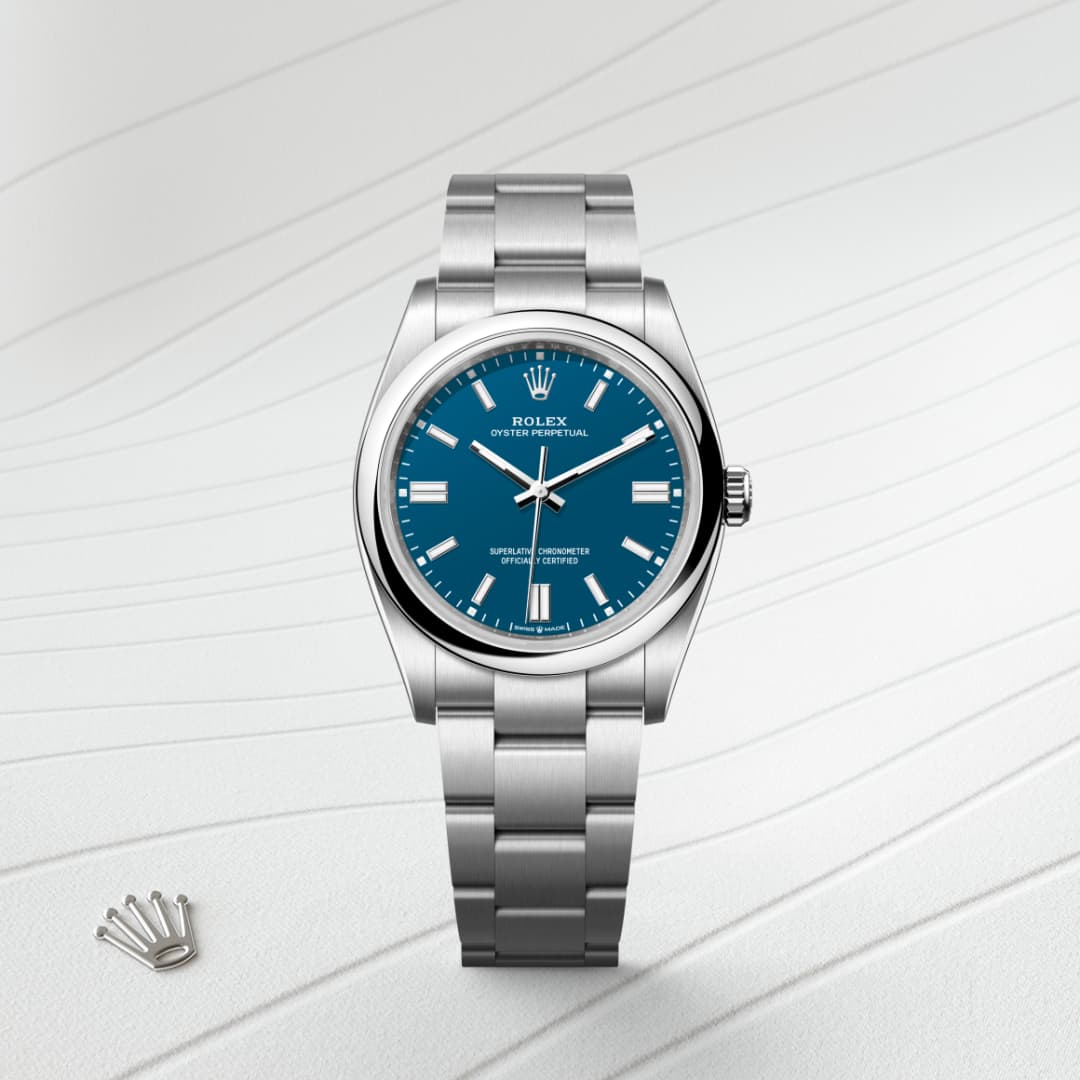 A silver Rolex watch with a blue dial is displayed on a textured white surface beside a small crown emblem.