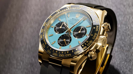 Gold Rolex chronograph watch with a light blue face and black subdials, displayed on a dark surface.