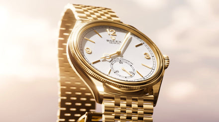 A gold Rolex wristwatch with a white face and gold bracelet, shown close-up against a soft, blurred background.