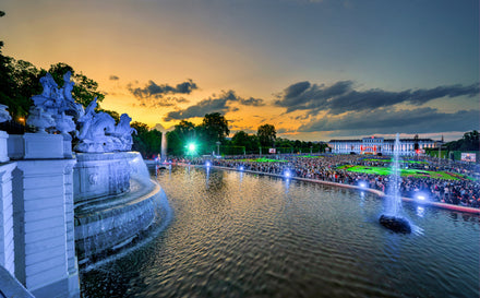 Sunset over a large fountain and crowded park with a grand palace in the background, lights illuminating the scene.