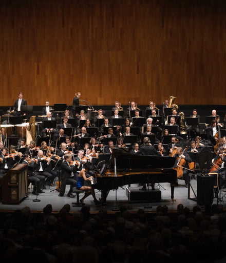 A symphony orchestra performs on stage with a pianist in front of a large seated audience.