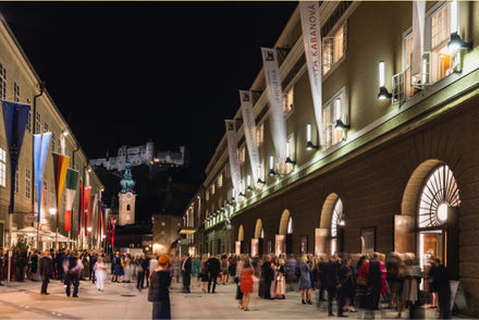 Crowds gather outside a brightly lit building at night, with flags and a castle visible in the background.