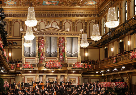 An orchestra performs on a grand, ornate stage decorated with pink flowers and large chandeliers.