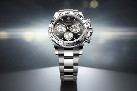 A silver Rolex Daytona watch with a black and silver dial displayed against a blurred, radiant background.