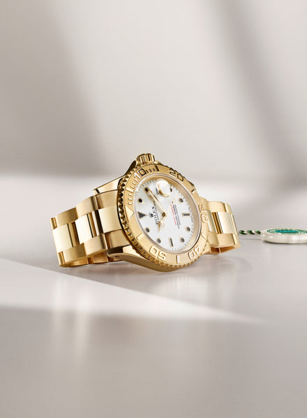 Gold luxury wristwatch with a white dial and metal bracelet, displayed on a light surface with a tag nearby.