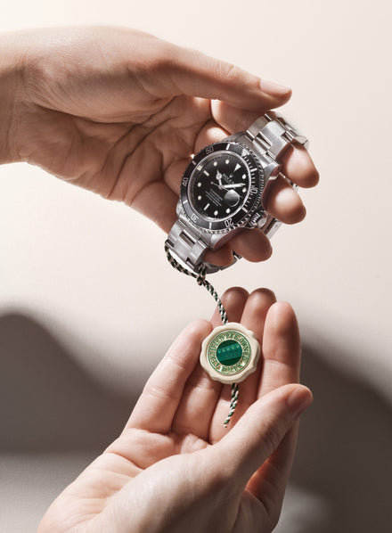A person holds a silver wristwatch and a green and white Rolex certified tag in their hands.