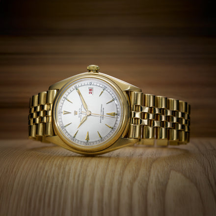 A gold wristwatch with a white dial and date display, placed on a wooden surface with a wooden background.