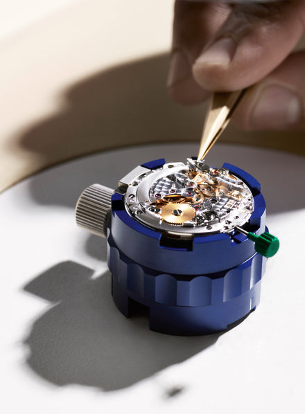 A hand uses tweezers to adjust the gears inside a mechanical watch movement on a blue holder.