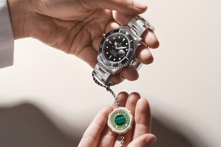 A hand holds a silver Rolex watch while another hand holds a green and white Rolex certification tag.